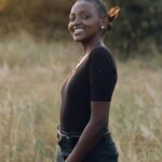 A smiling woman wearing a black top stands in a grassy field during sunset, embodying natural beauty and joy.